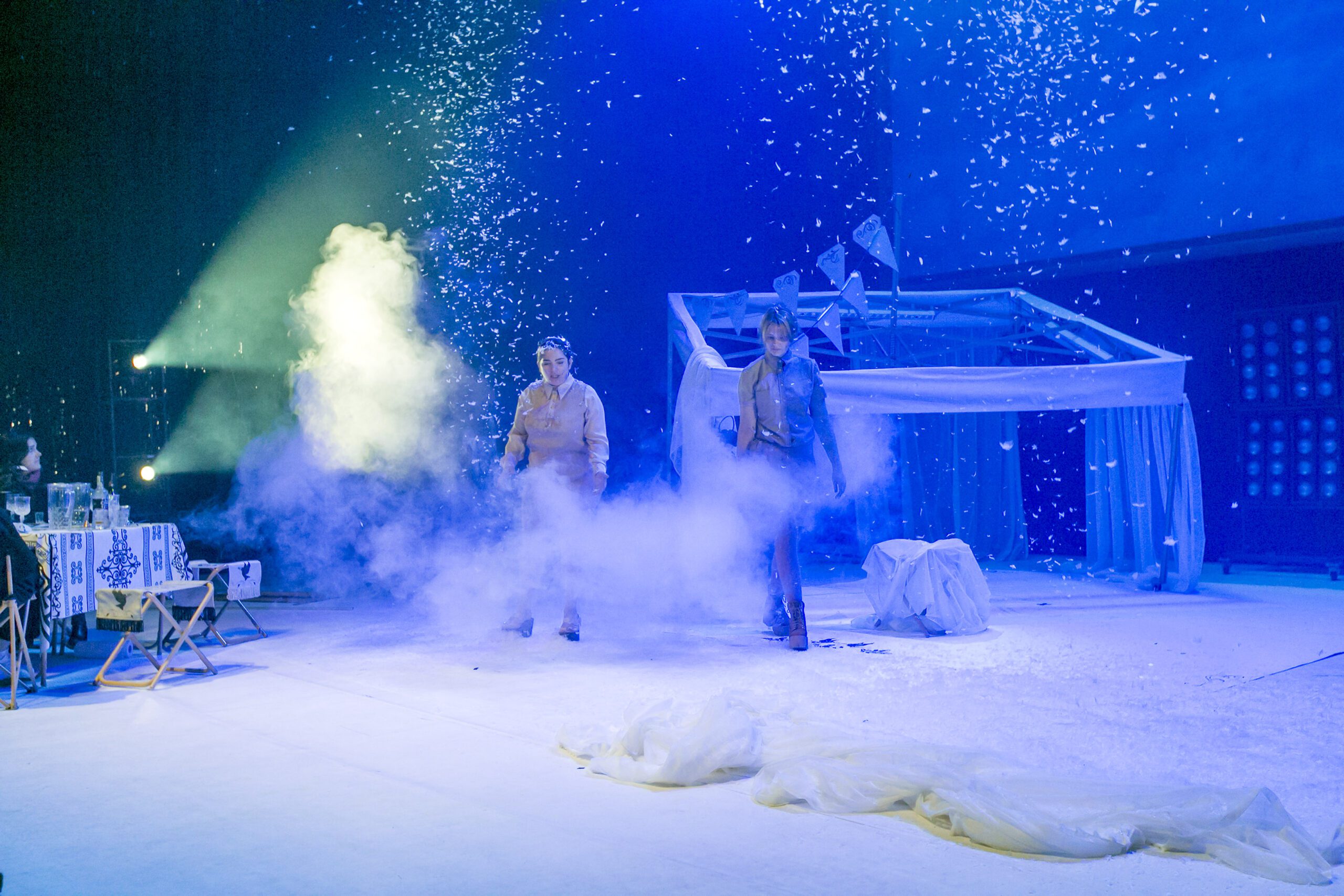 Two actors perform on a brightly lit stage surrounded by artificial snow, smoke, and blue lighting. A structure resembling a house frame and a table with chairs are also part of the scene.