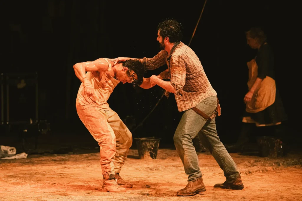 A man in a plaid shirt grabs the head of another man covered in mud during a stage performance.
