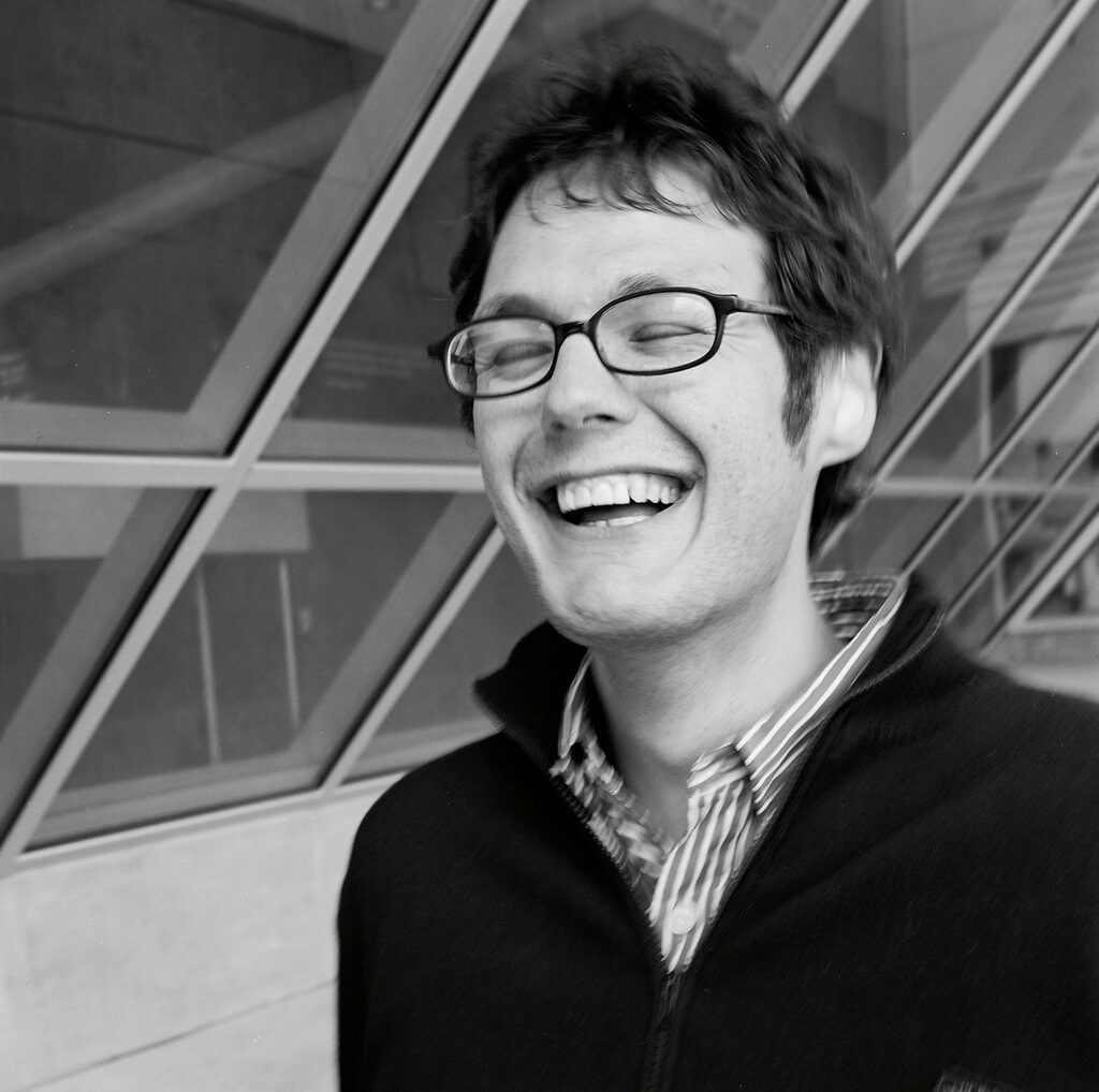 A person with glasses laughing with their eyes closed, standing in front of a glass building.