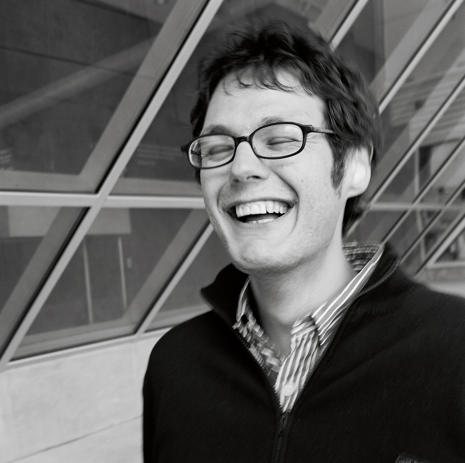 A person with glasses laughing with their eyes closed, standing in front of a glass building.